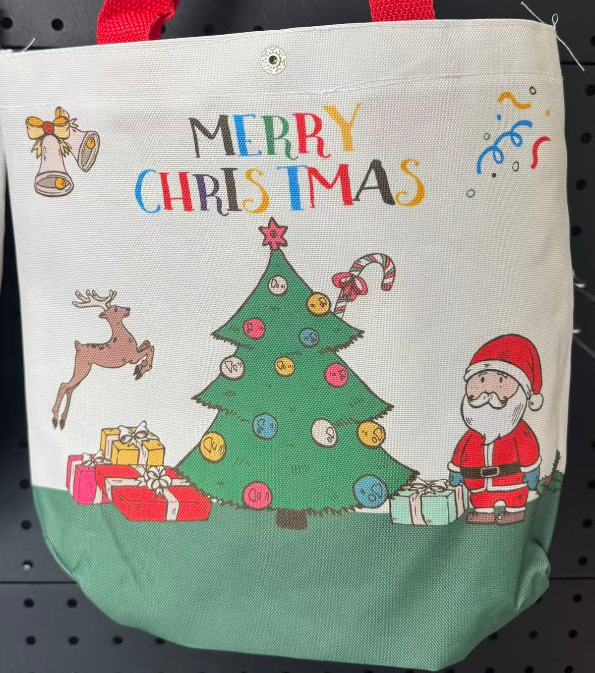 A canvas Christmas tote bag with a cream upper section and a sage green lower section, featuring a colorful 'MERRY CHRISTMAS' print with Santa, a reindeer, a Christmas tree, and presents on a black pegboard background.