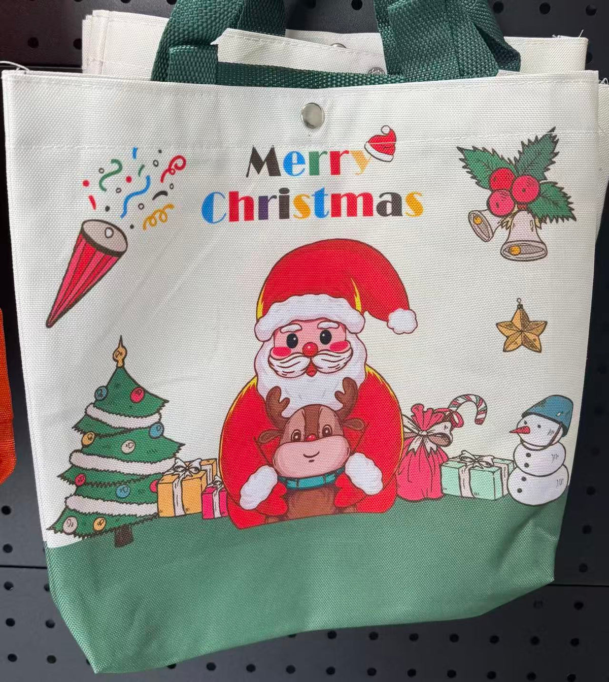 A canvas tote bag with a green bottom panel and green handles, featuring a colorful Christmas print with Santa, a reindeer, a tree, gifts, and the words 'Merry Christmas' on a white upper section.